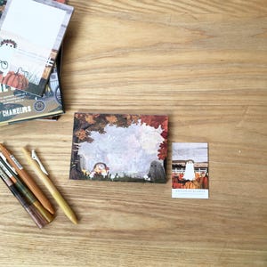 May include: A collection of stationery items, including notepads, pens, and a small card, arranged on a wooden surface. The notepads feature illustrations of ghosts and pumpkins. The pens are in shades of brown and gold.