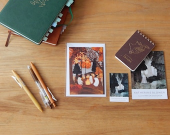 May include: A collection of stationery items including a green notebook, a brown spiral notepad with Totoro text, three pens, a postcard featuring ghosts around a campfire, and two business cards with the text Katherine Blower Illustrator/Designer.
