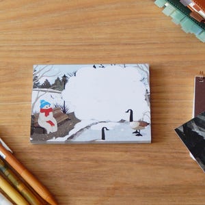 May include: A winter-themed notepad featuring a snowman wearing a blue hat and scarf, sitting on a bench. The illustration includes geese, trees, and a snowy landscape. The notepad is surrounded by pens, notebooks, and a card on a wooden surface.