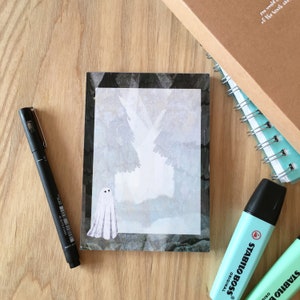 Puede incluir: Un fantasma blanco con dos ojos negros está de pie frente a un fondo de acuarela gris y negro. El fondo parece ser una escena de bosque.