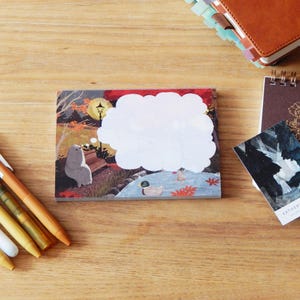 May include: A stack of illustrated sticky notes with a cloud-shaped blank space, featuring a park scene with a bear. Also, a small notebook, a card, and several pens are on a wooden surface.