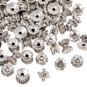 May include: A collection of silver-colored metal bead caps in various shapes, including floral, bell, and butterfly designs. The bead caps have a distressed, antique finish and are scattered across a white surface.