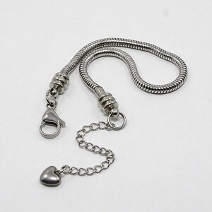 May include: A silver-colored bracelet chain with a heart-shaped charm. The chain has a snake-like design and includes a lobster clasp and an extender chain. The heart charm is small and smooth, and the chain is on a white background.