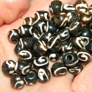 May include: A collection of dark brown beads with white, swirling patterns. Each bead is round with a hole through the center, suggesting they are for jewelry making or crafts. The beads are held in a hand.
