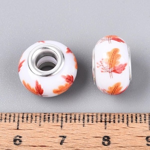 May include: Two white round beads with orange and red leaf patterns, each featuring a silver-toned metal core. A wooden ruler is positioned below the beads, showing they measure approximately 0.5 inches in diameter and height.