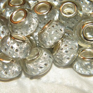 May include: A close-up of a pile of silver and gold-rimmed glass beads with a sparkly, textured surface. The beads are round and have a hole in the center for threading.