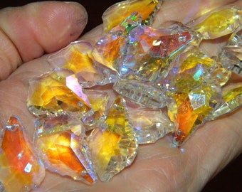 May include: A collection of clear, faceted crystal pendants featuring an iridescent finish that reflects vibrant orange, yellow, and rainbow hues. Each individual crystal piece has a small hole at the top for hanging, showcasing a geometric shape.