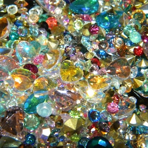 May include: A close-up of a variety of colorful, iridescent, and clear glass rhinestones. The rhinestones are in various shapes, including hearts, ovals, and rounds. The rhinestones are scattered on a black surface.