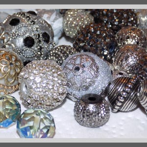 May include: A collection of assorted craft beads in various textures and finishes, including silver glitter spheres, dark metallic faceted beads, clear iridescent crystals, and round beads featuring intricate gold-toned filigree metal overlays.