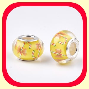 May include: Two yellow glass beads with a silver metal core. The beads are decorated with a pattern of red and white candy canes and orange bows.