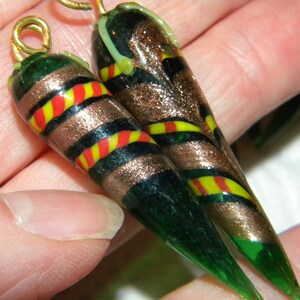 May include: Two green glass teardrop-shaped beads with gold and black stripes, red and yellow accents, and gold accents. The beads have a loop at the top for hanging.