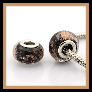 May include: Two black and gold glass beads with silver metal accents. The beads have a speckled pattern and are designed to fit on a charm bracelet.