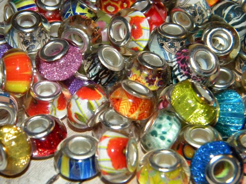 May include: A collection of colorful glass beads with silver-toned metal accents. The beads feature various designs, including floral patterns, glitter, and abstract art. The beads are in shades of red, orange, yellow, green, blue, and purple.