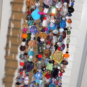 May include: Strands of colorful beads in various shapes and sizes, including clear, blue, red, and orange. The beads are strung together, creating a decorative piece. The beads are hanging in front of a white shutter.