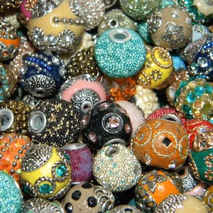 May include: A collection of assorted round beads featuring various textures, colours, and metallic accents. The beads include designs with embedded rhinestones, intricate silver-toned filigree patterns, and textured surfaces in shades of blue, orange, yellow, black, and beige.