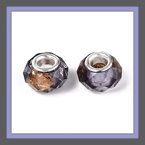 May include: Two faceted, purple and gold-toned glass beads with silver-colored metal accents. The beads have a geometric design and are displayed against a white background. These beads are suitable for jewelry making.