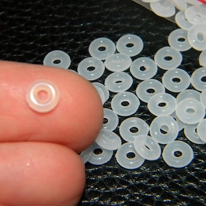 May include: A large quantity of small, white, plastic rings. The rings are approximately 1.2 cm in diameter and have a small hole in the centre. The rings are arranged in a random pattern on a black surface.