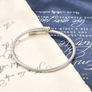 May include: A silver-toned cable-style bangle bracelet featuring a twisted metal wire design. The bracelet has a circular shape with two rounded, smooth metal end caps that meet at the opening, creating a clean and simple aesthetic for the wrist.