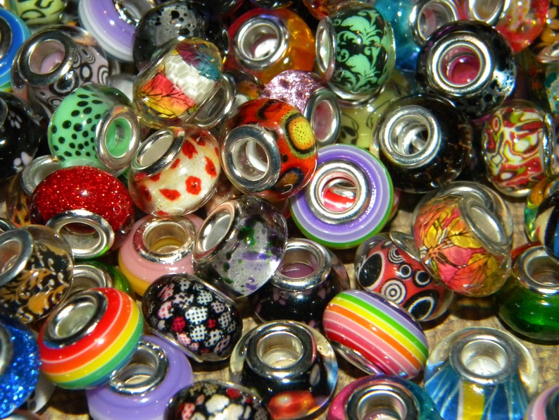 May include: A collection of colorful glass beads with silver-toned metal cores. The beads feature various designs, including floral patterns, polka dots, and rainbow stripes. The beads are in shades of red, green, blue, purple, and orange.