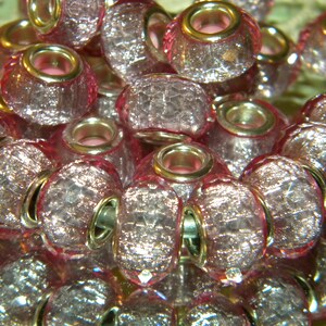 May include: A collection of pink, faceted glass beads with silver-colored metal accents. The beads have a textured, crackled appearance and are arranged closely together, reflecting light. The beads have a hole through the center for stringing.