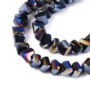 May include: Close-up of two strands of faceted, dark gray beads with iridescent color shifts. The beads have a geometric, angular shape, reflecting light with hues of blue, purple, and gold. The beads are strung together, suggesting jewelry making.