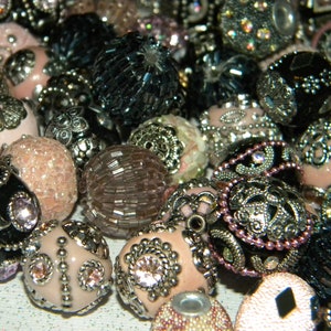 May include: A collection of decorative beads in various colors and styles, including pink, black, and silver. The beads are made of glass, metal, and other materials. Some beads have intricate designs and patterns.