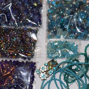 May include: A collection of assorted craft beads in various shades of blue, purple, and teal. The set includes multiple clear plastic bags filled with loose beads, strands of turquoise beads, and a decorative metal floral brooch with blue stones.
