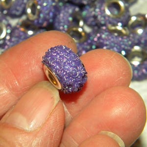 May include: A close-up of a purple bead with a sparkly, textured surface. The bead has a gold-colored metal core. Other similar beads are visible in the background, out of focus. The beads are likely for jewelry making.