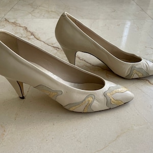 May include: A pair of off-white leather pumps with a low heel. The toe of each shoe features a decorative design in shades of gold and gray. The shoes are set on a light-colored surface.