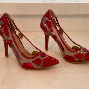 May include: A pair of red patent leather high heels with clear panels. The shoes have a pointed toe and a stiletto heel. The red sections have a unique pattern. The shoes are displayed on a white surface.