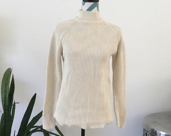 May include: Cream-colored ribbed knit turtleneck sweater displayed on a mannequin. The long-sleeved sweater has a high neckline and a classic, simple design. The sweater is made of a soft, textured material, perfect for cooler weather.