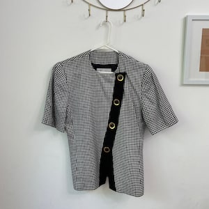 May include: A black and white houndstooth pattern short-sleeve top. The top features a black vertical panel with gold-colored circular accents. The top is hanging on a white hanger against a white wall.