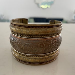 Puede incluir: Un brazalete ancho apilado con un acabado de lat&oacute;n antiguo. El brazalete presenta patrones en relieve detallados, incluido un dise&ntilde;o de elefante en la banda central. El brazalete de metal mide aproximadamente 5 cm de ancho.