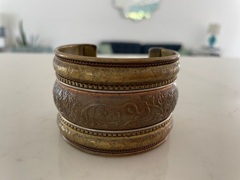 Puede incluir: Un brazalete ancho apilado con un acabado de lat&oacute;n antiguo. El brazalete presenta patrones en relieve detallados, incluido un dise&ntilde;o de elefante en la banda central. El brazalete de metal mide aproximadamente 5 cm de ancho.