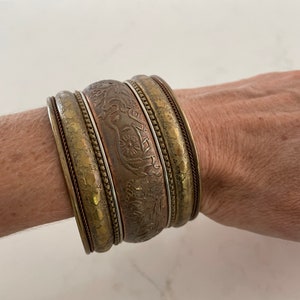 Puede incluir: Un conjunto de brazaletes antiguos de lat&oacute;n y cobre. Los brazaletes presentan intrincados dise&ntilde;os florales y geom&eacute;tricos en relieve. Los brazaletes est&aacute;n apilados en una mu&ntilde;eca, mostrando sus diferentes anchos y texturas. La paleta de colores incluye tonos dorados, bronce y cobre.