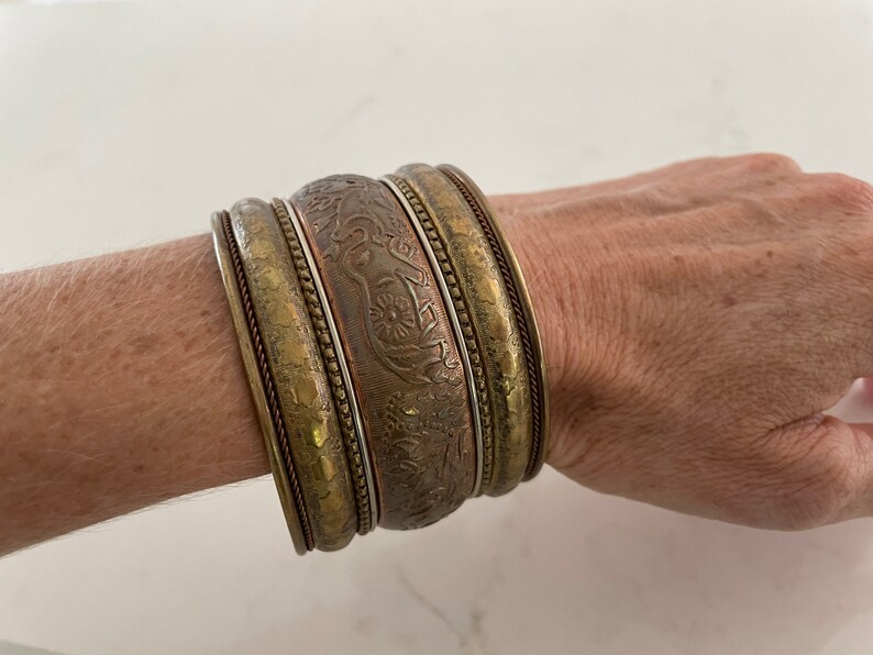 Puede incluir: Un conjunto de brazaletes antiguos de lat&oacute;n y cobre. Los brazaletes presentan intrincados dise&ntilde;os florales y geom&eacute;tricos en relieve. Los brazaletes est&aacute;n apilados en una mu&ntilde;eca, mostrando sus diferentes anchos y texturas. La paleta de colores incluye tonos dorados, bronce y cobre.