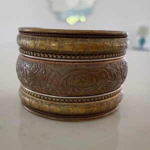 Puede incluir: Conjunto apilado de cuatro pulseras met&aacute;licas. Las pulseras superior e inferior son de color dorado con un patr&oacute;n texturizado. La pulsera central es de color cobre con un dise&ntilde;o en relieve de elefante y flores.