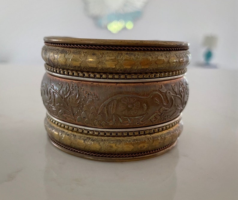 Puede incluir: Conjunto apilado de cuatro pulseras met&aacute;licas. Las pulseras superior e inferior son de color dorado con un patr&oacute;n texturizado. La pulsera central es de color cobre con un dise&ntilde;o en relieve de elefante y flores.