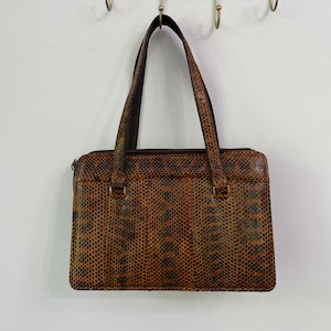 May include: A brown and black snakeskin-patterned handbag with two handles. The rectangular bag has a zip closure and is hanging from a gold-coloured hook. The bag's pattern resembles reptile skin.