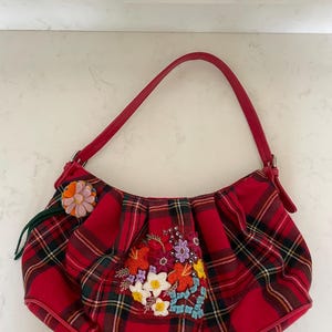 May include: Red plaid shoulder bag with a red leather strap. The bag features an embroidered floral design with colorful accents. A small, decorative flower is attached to the side. The bag is a stylish accessory.