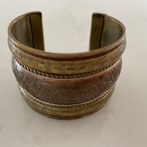 Puede incluir: Pulsera de pu&ntilde;o ancha y abierta en tonos oro antiguo y bronce. La pulsera presenta patrones en relieve detallados, incluyendo motivos florales y animales. El brazalete de metal mide aproximadamente 5 cm de ancho y tiene una superficie texturizada.