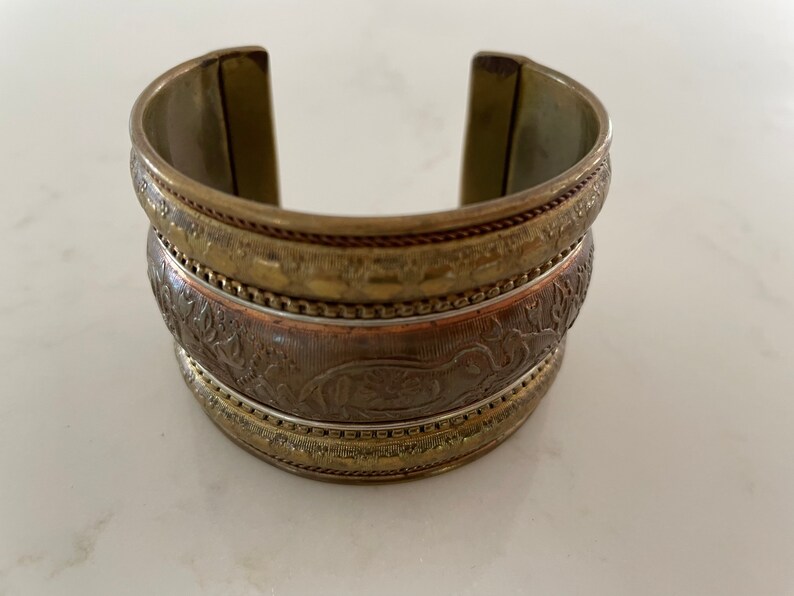 Puede incluir: Pulsera de pu&ntilde;o ancha y abierta en tonos oro antiguo y bronce. La pulsera presenta patrones en relieve detallados, incluyendo motivos florales y animales. El brazalete de metal mide aproximadamente 5 cm de ancho y tiene una superficie texturizada.