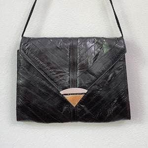 May include: Black leather crossbody bag with a geometric design. The bag features a flap closure and a decorative metal accent. The strap is a thin, braided cord. The bag is rectangular and has a textured surface.