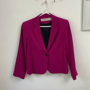May include: A magenta blazer with a black grid pattern, single-button closure, and a black lining. The blazer is hanging on a white hanger against a white wall. The label reads "Little Robin".