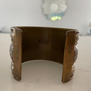 Puede incluir: Pulsera ancha de lat&oacute;n con detalles plateados ornamentados. La pulsera tiene forma rectangular con patrones decorativos y una est&eacute;tica vintage. El metal tiene un aspecto c&aacute;lido y envejecido.