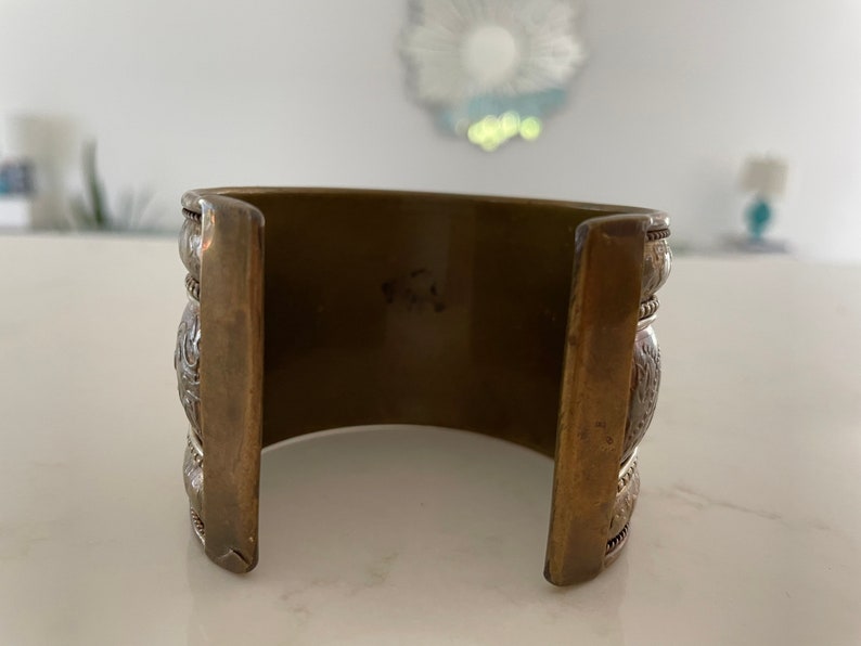 Puede incluir: Pulsera ancha de lat&oacute;n con detalles plateados ornamentados. La pulsera tiene forma rectangular con patrones decorativos y una est&eacute;tica vintage. El metal tiene un aspecto c&aacute;lido y envejecido.