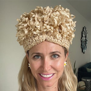 May include: A beige, woven hat with a textured, ribbon-like design on top. The hat has a rounded shape and appears to be made of natural fibers. The hat is worn by a person with blonde hair and daisy earrings.