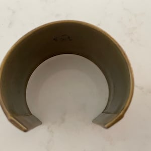 Puede incluir: Un brazalete de pu&ntilde;o ancho y abierto con un acabado de lat&oacute;n cepillado. El interior es de color gris apagado. El brazalete tiene un dise&ntilde;o simple y minimalista, con una ligera curva para adaptarse a la mu&ntilde;eca. El metal tiene un aspecto ligeramente envejecido.