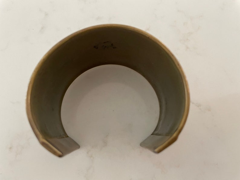 Puede incluir: Un brazalete de pu&ntilde;o ancho y abierto con un acabado de lat&oacute;n cepillado. El interior es de color gris apagado. El brazalete tiene un dise&ntilde;o simple y minimalista, con una ligera curva para adaptarse a la mu&ntilde;eca. El metal tiene un aspecto ligeramente envejecido.