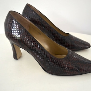 May include: A pair of brown and black snakeskin-patterned pumps with a pointed toe and a mid-height heel. The shoes are displayed on a white surface, showcasing their textured design and classic style.